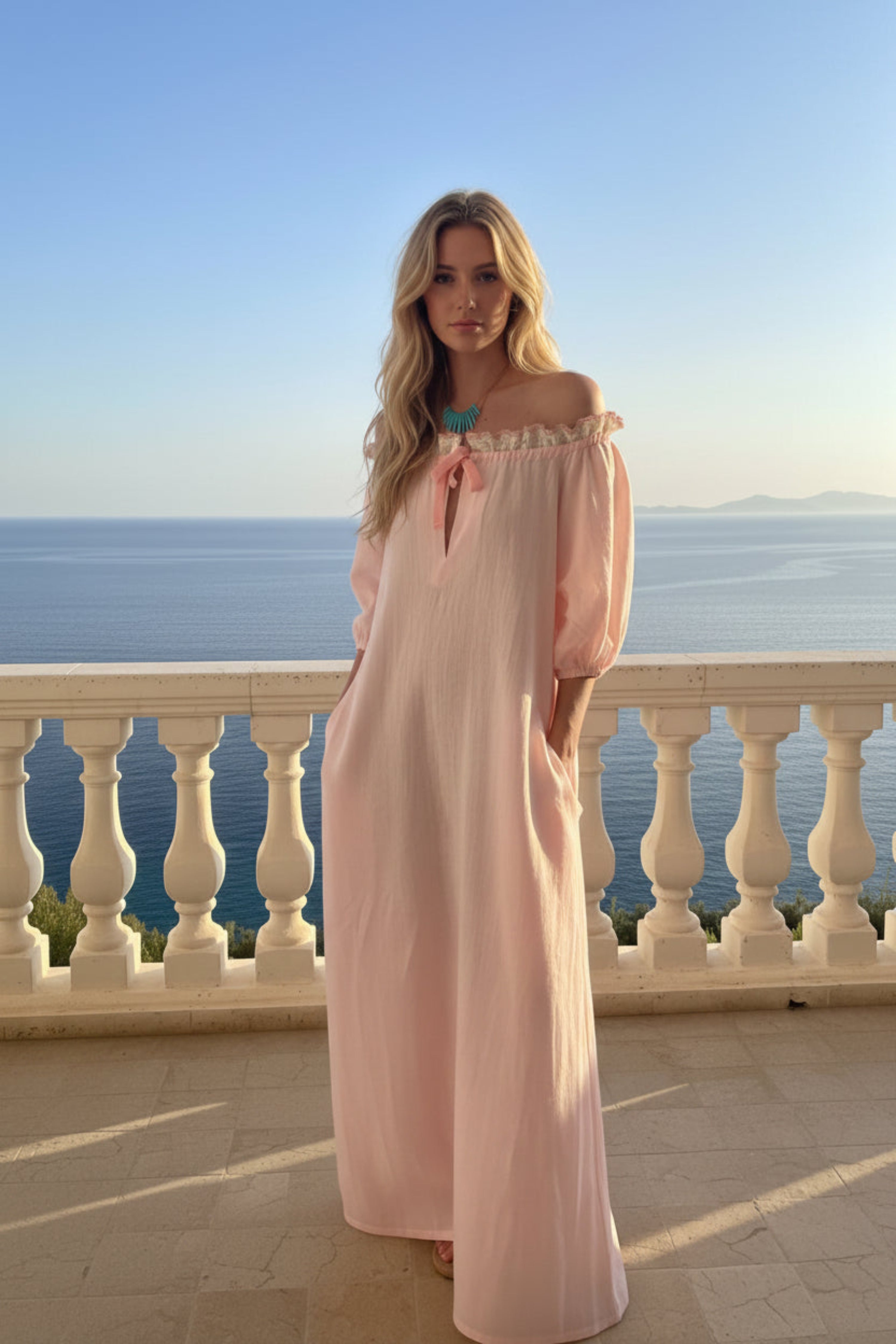 A woman wearing a light pink off-the-shoulder maxi dress with a tie-neck detail, standing on a sunlit terrace with a white balustrade and the ocean in the background.